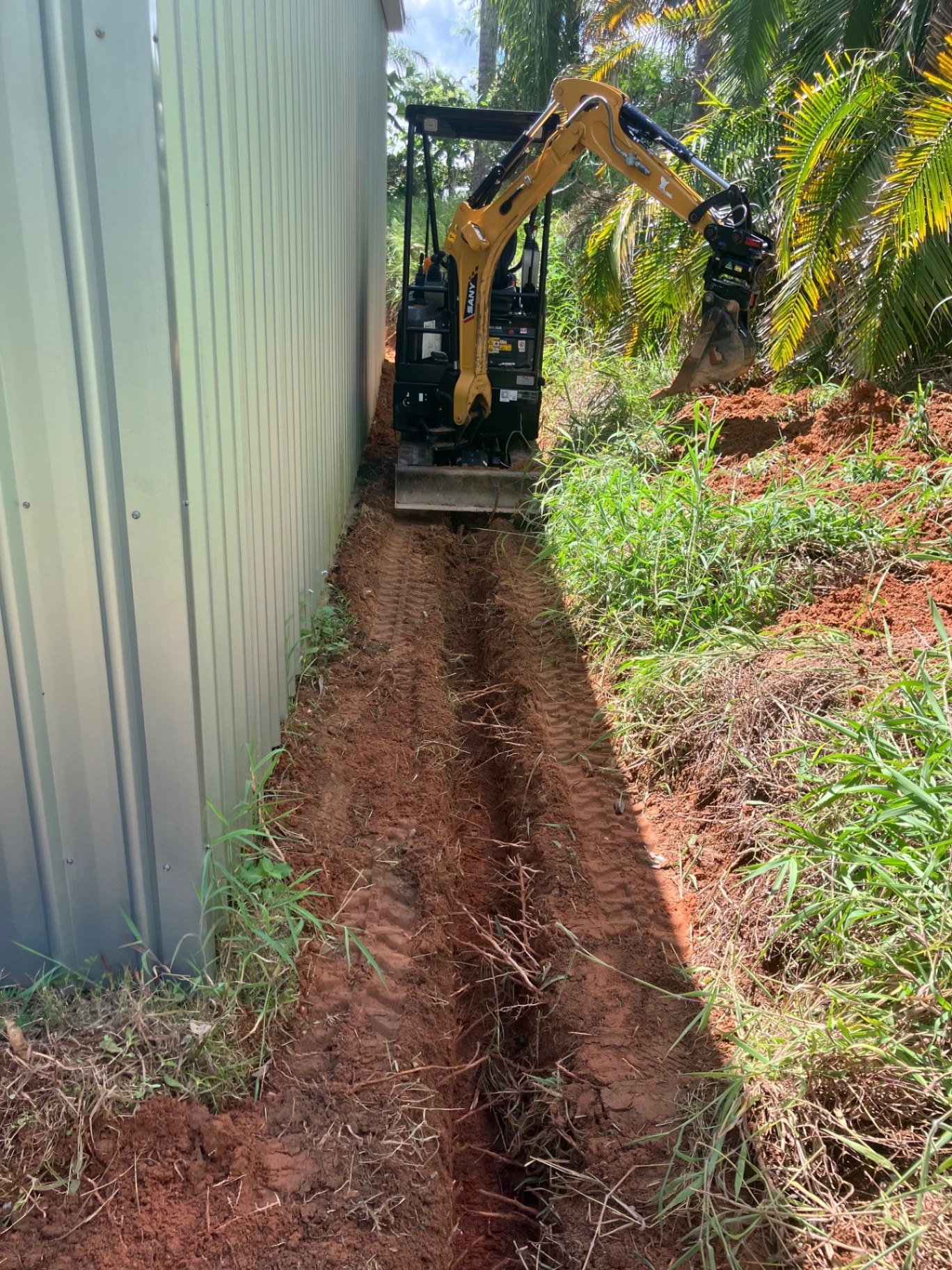 Mini excavator digging a narrow trench between a shed wall and tropical garden